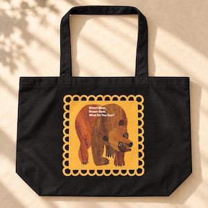 May include: A black tote bag featuring a central yellow square with a scalloped border. Inside the square is a brown bear illustration and the white text: Brown Bear, Brown Bear, What Do You See? The bag has two black fabric handles at the top.