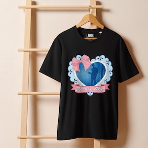 Puede incluir: Camiseta negra con un diseño en forma de corazón. Presenta la silueta de una persona, un lazo rosa y el texto "Dream Guy" en una pancarta. El diseño está enmarcado por un borde festoneado azul claro.