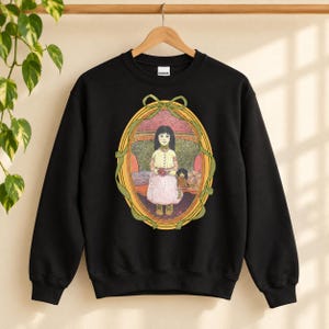 Puede incluir: Una sudadera negra de cuello redondo con un estampado gráfico central de una persona con cabello largo y oscuro que viste un vestido claro y sostiene una muñeca pequeña. La ilustración está enmarcada por un borde ovalado con un lazo verde.