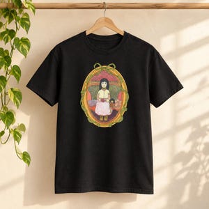 Puede incluir: Una camiseta negra de manga corta con un gráfico central de una persona de pelo oscuro con una blusa amarilla y una falda rosa, dentro de un marco ovalado dorado adornado con un lazo verde en la parte superior sobre un fondo rojo.