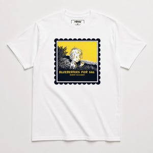 Op de afbeelding: Een wit t-shirt met korte mouwen en een centrale afbeelding van een kind in een veld met de tekst Blueberries for Sal en Robert McCloskey. Het ontwerp is omlijst door een donkerblauwe geschulpte rand op een gele achtergrond.