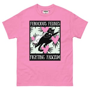 Könnte beinhalten: Rosafarbenes T-Shirt mit einer Grafik, die eine schwarze Katze mit weißem Umhang zeigt, umgeben von rosa Herzen und dem Text "FEROCIOUS FELINES FIGHTING FASCISM". Das T-Shirt hat einen Rundhalsausschnitt und kurze Ärmel.