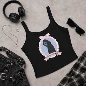 Puede incluir: Un top corto negro sin mangas con un gráfico de una figura encapuchada en un marco ovalado azul claro con un lazo y corazones rosas. El texto HE GOT ME FEELING WOOZY está impreso en una cinta rosa en la parte inferior del diseño.