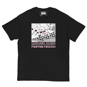 Könnte beinhalten: Schwarzes T-Shirt mit einem Cartoon-Katzendesign, einer rosa Schleife und Noten. Der Text "FEROCIOUS FELINES FIGHTING FASCISM" ist rosa. Die Katze befindet sich vor einem karierten und rasterförmigen Hintergrund.