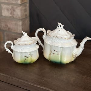 May include: A vintage tea set comprising a teapot and sugar bowl. The white porcelain set has gold trim and a green gradient at the base. The pieces are decorated with delicate leaf designs, giving a classic, elegant appearance.