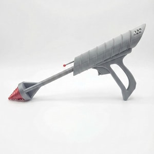 May include: A gray ray gun with a red drill-like tip and a red antenna. The gun has a ribbed barrel and a handle. The top of the gun has several small holes. The ray gun is a prop or toy.