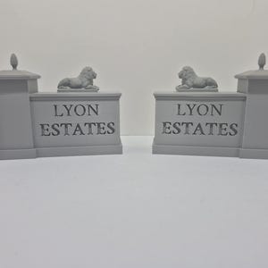 以下が含まれることがあります： ライオンの像が上に乗った、2つの灰色のミニチュア門柱。各柱の前面には「LYON ESTATES」の文字が刻まれています。柱は明るい灰色で、シンプルでクラシックなデザインです。