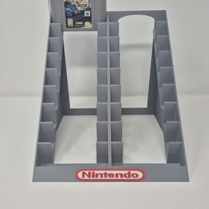 以下が含まれることがあります： ビデオゲームカートリッジ用のグレーのプラスチック製収納ラックで、2列の垂直スロットがあります。1つのスロットには、NINTENDO 64コンソール用のNASCAR 2000ゲームカートリッジが入っています。ラックのベースには、赤と白の任天堂のロゴエンブレムが付いています。