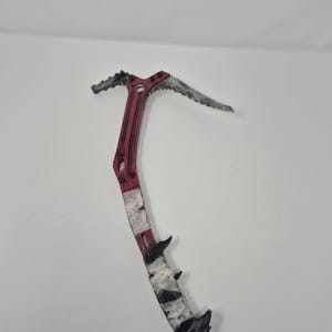 May include: A red and silver ice axe with a curved blade and a spiked end. The handle is wrapped with white tape. The axe is designed for climbing and mountaineering.