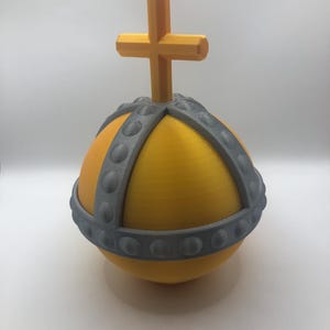May include: A 3D-printed yellow orb with a gray band and a cross on top. The orb has a textured surface and a decorative gray band around the middle. The cross is also yellow and sits atop the orb.