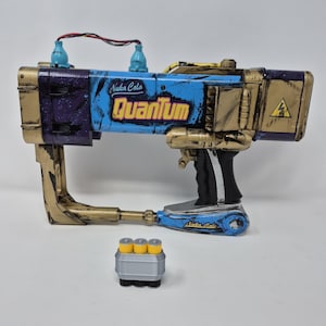 May include: A prop gun with a blue and gold color scheme, featuring the text "Nuka Cola Quantum." The gun has a handle, a barrel, and two blue bottle-shaped attachments. A separate cartridge with three yellow cylinders is also present.