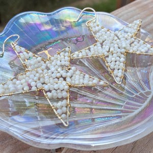 May include: A pair of star-shaped earrings with gold-colored wire frames and filled with small, white beads. The earrings are displayed on a clear, iridescent glass dish. The earrings have a hook closure.