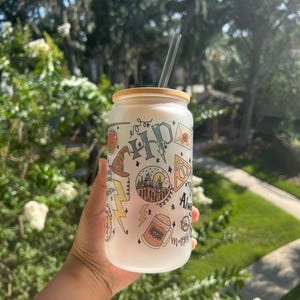 May include: A frosted glass can-shaped tumbler with a bamboo lid and a clear straw. The tumbler is decorated with colorful illustrations and text, including a lightning bolt, a wizard's hat, and the word "Always".