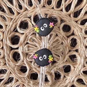 Peut inclure: Deux bâtonnets à cocktail noirs avec des personnages de dessins animés, chacun avec des yeux blancs et des étoiles colorées. Ils sont posés sur un dessous de plat circulaire en fibres naturelles. Le fond est sombre.