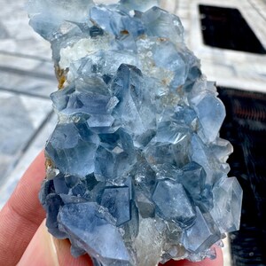 May include: A cluster of blue celestite crystals. The crystals have a translucent appearance and a range of blue hues, from light to deep. The crystals are clustered together, forming a rough, irregular shape. The overall effect is one of natural beauty and geological interest.
