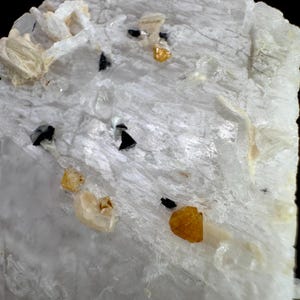 Puede incluir: Primer plano de un espécimen mineral con una estructura cristalina blanca. El espécimen presenta pequeñas inclusiones negras y amarillo dorado dispersas. La superficie tiene un aspecto texturizado, con formaciones cristalinas visibles y variaciones de color.