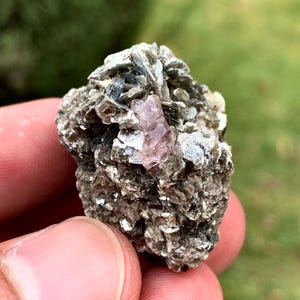 May include: A raw mineral specimen featuring a cluster of metallic, greyish-brown mica flakes surrounding a single, translucent, pale pink crystal formation embedded in the centre of the rough, textured rock structure.