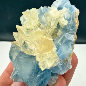 May include: A raw mineral specimen featuring a cluster of translucent, light blue crystals intermixed with a prominent formation of pale yellow, sharp-edged crystals. The structure displays varied geometric facets and a natural, rugged texture.