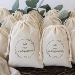 May include: A collection of beige drawstring bags. Each bag features a circular design with a floral wreath, the text "Add your text/logo/photo," and a heart. The bags are arranged together, with some greenery in the background.