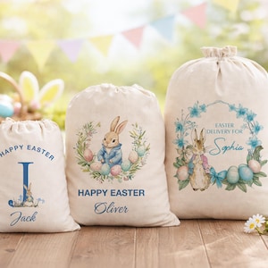 May include: Three off-white drawstring canvas bags featuring Easter-themed illustrations. The bags display the names Jack, Oliver, and Sophia, along with printed text including Happy Easter and Easter Delivery For, accompanied by rabbit and egg graphics.