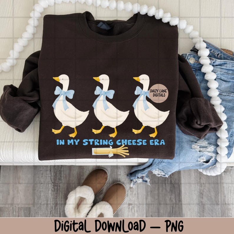 May include: A dark brown sweatshirt with a design of three cartoon geese wearing blue bows. The text "IN MY STRING CHEESE ERA" is below the geese, with a string cheese graphic. The words "Digital Download - PNG" are at the bottom.