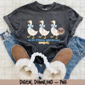 May include: Dark grey t-shirt with a graphic of three white geese wearing blue bows, with the text "IN MY STRING CHEESE ERA" and a string cheese illustration. The shirt is styled with blue jeans and fluffy slippers.