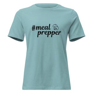 May include: A light blue t-shirt with short sleeves. The shirt features the text "#meal prepper" in black, along with a small graphic of broccoli. The shirt is made of a soft material.