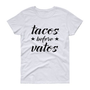 May include: White t-shirt with the phrase "tacos before vatos" in black script. The text is centered and includes stars on either side of the word "before". The shirt has a crew neck and short sleeves.