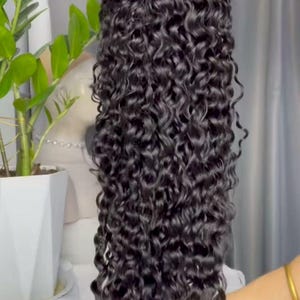 May include: A long, dark curly wig with a natural look. The hair is a deep black shade and has tight, bouncy curls. The wig is held up, showing its length and volume. The background includes a plant and a white surface.