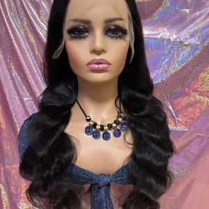 May include: A mannequin head with long, wavy black hair, styled into loose curls. The mannequin wears a blue top and a necklace with blue and gold accents. The face has dramatic makeup, including dark eye shadow and long eyelashes.