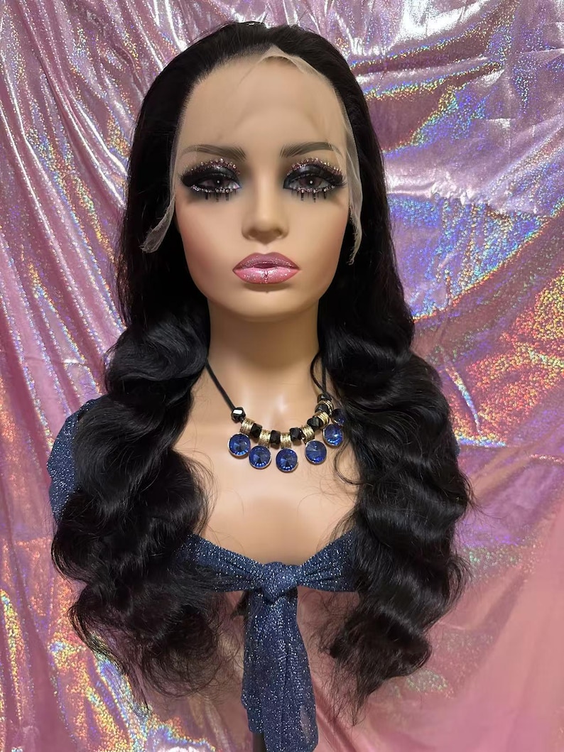 May include: A mannequin head with long, wavy black hair, styled into loose curls. The mannequin wears a blue top and a necklace with blue and gold accents. The face has dramatic makeup, including dark eye shadow and long eyelashes.