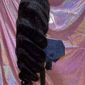 May include: A long, wavy black wig styled with voluminous curls. The wig is displayed on a mannequin head with a denim-like fabric covering the neck area. The background is a shiny, pink, iridescent material.