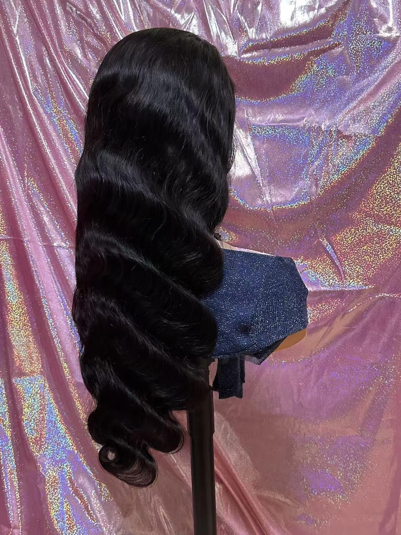 May include: A long, wavy black wig styled with voluminous curls. The wig is displayed on a mannequin head with a denim-like fabric covering the neck area. The background is a shiny, pink, iridescent material.