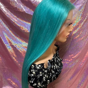 May include: A turquoise wig with long, straight hair is displayed on a mannequin head. The wig has a lace front and is styled with a side part. The mannequin is wearing a black top with a white floral pattern. The background is a shiny, pink surface.