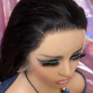 May include: A black lace front wig styled on a mannequin head. The wig features long, dark hair with a natural hairline. The mannequin has dramatic eye makeup with long eyelashes and a blue and black necklace.
