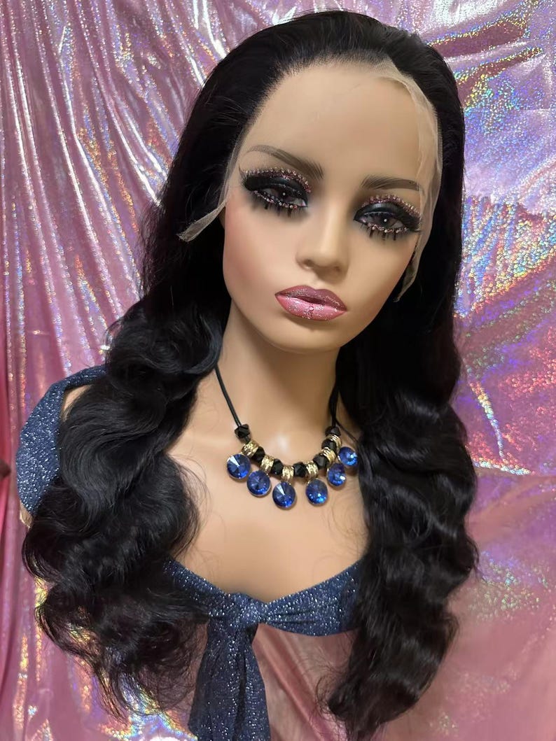 May include: A mannequin head with long, wavy black hair and a lace front wig. The mannequin wears a blue and black beaded necklace with blue teardrop-shaped gems. The mannequin has dramatic eye makeup and pink lipstick.