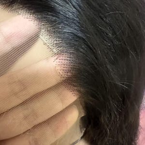 May include: Close-up of a black wig with a lace front. The wig features dark, wavy hair and a delicate, sheer lace base. The lace is a light skin tone, and the hair is neatly styled.