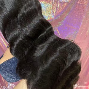 May include: A bundle of long, wavy, black hair. The hair is styled in loose waves, with a natural shine. The hair is held in place, showing the texture and volume of the hair. The background is a pink and purple iridescent material.