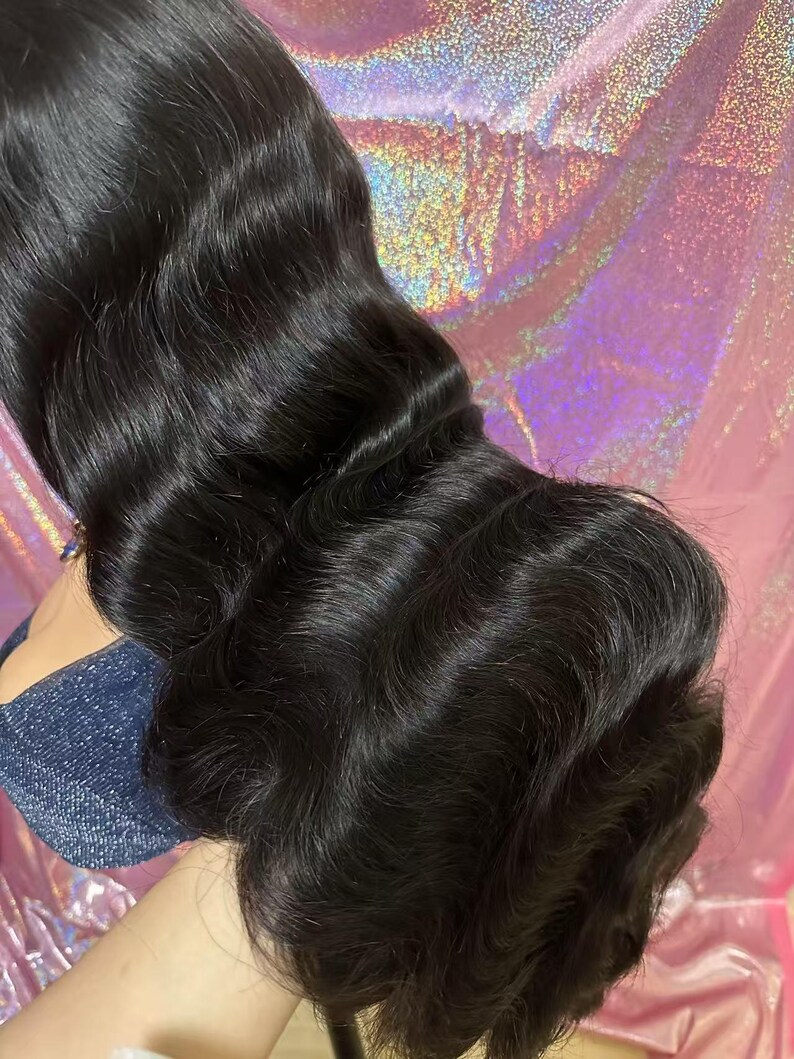 May include: A bundle of long, wavy, black hair. The hair is styled in loose waves, with a natural shine. The hair is held in place, showing the texture and volume of the hair. The background is a pink and purple iridescent material.