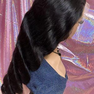 May include: A long, wavy black wig styled on a mannequin head. The wig features a lace front and is styled with loose curls. The mannequin is wearing a blue denim-like top. The background is a pink, iridescent fabric.