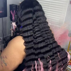 May include: Long, wavy black hair extensions with a crimped texture. The hair flows down the back, with a pinkish hue at the ends. The person is holding a phone.