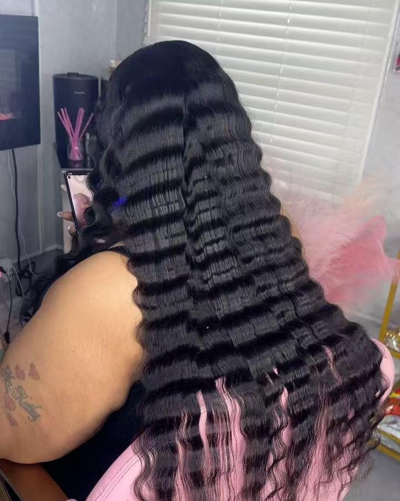 May include: Long, wavy black hair extensions with a crimped texture. The hair flows down the back, with a pinkish hue at the ends. The person is holding a phone.