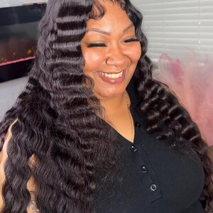 May include: A woman with long, wavy, dark brown hair. The hair is styled with a deep part and loose waves. She is wearing a black top and smiling. The image is well-lit, with a focus on the hair and the woman's expression.
