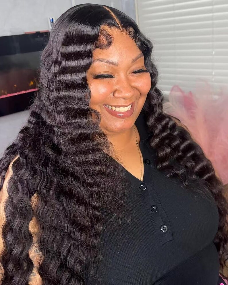 May include: A woman with long, wavy, dark brown hair. The hair is styled with a deep part and loose waves. She is wearing a black top and smiling. The image is well-lit, with a focus on the hair and the woman's expression.