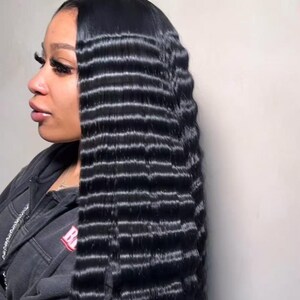 May include: A side view of a person with long, crimped black hair. The hair is styled with a deep side parting and falls down the back in a wavy pattern. The person is wearing a dark grey hooded sweatshirt.