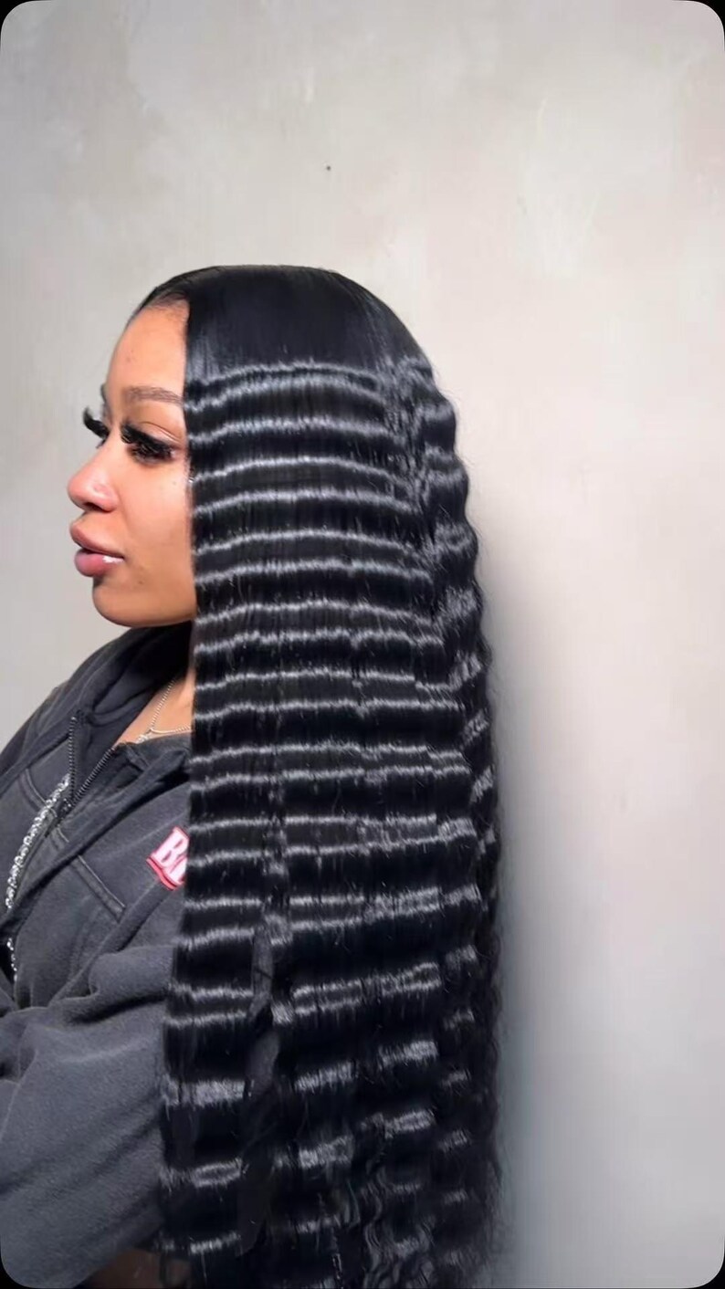 May include: A side view of a person with long, crimped black hair. The hair is styled with a deep side parting and falls down the back in a wavy pattern. The person is wearing a dark grey hooded sweatshirt.