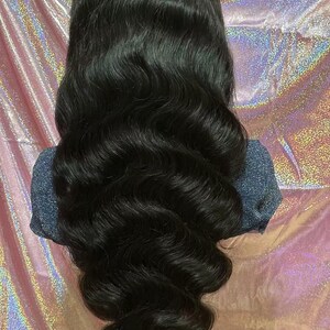May include: A long, wavy black wig with a natural-looking hairline. The hair is styled in loose, voluminous waves, cascading down. The wig is displayed against a pink and silver iridescent background.