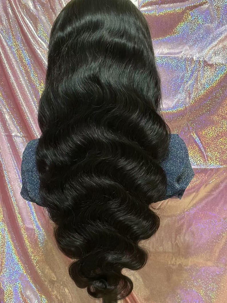 May include: A long, wavy black wig with a natural-looking hairline. The hair is styled in loose, voluminous waves, cascading down. The wig is displayed against a pink and silver iridescent background.