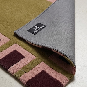 Pu&ograve; includere: Un tappeto rettangolare con un motivo geometrico di quadrati color verde oliva, rosa polvere e bordeaux scuro. L'angolo &egrave; ripiegato per rivelare un retro in tessuto grigio con un'etichetta nera che riporta la scritta M Majestic Rugs in bianco.