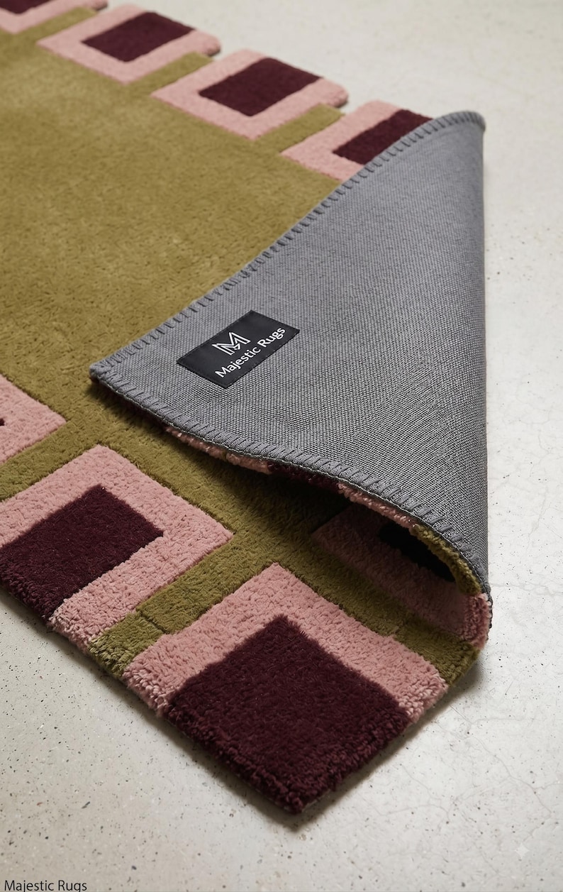 Pu&ograve; includere: Un tappeto rettangolare con un motivo geometrico di quadrati color verde oliva, rosa polvere e bordeaux scuro. L'angolo &egrave; ripiegato per rivelare un retro in tessuto grigio con un'etichetta nera che riporta la scritta M Majestic Rugs in bianco.